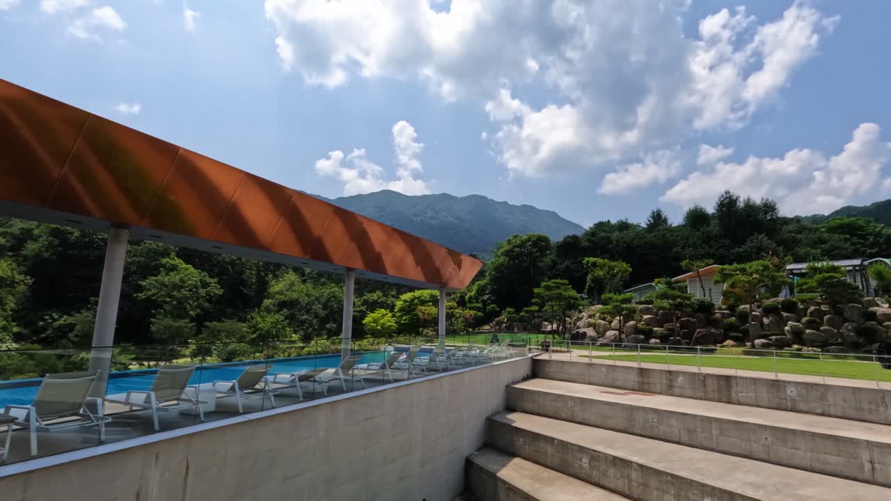 A timelapse captures beautiful moving clouds above a luxury resort swimming pool with a modern canopy and architectural concrete steps, set against a green mountain in Gapyeong