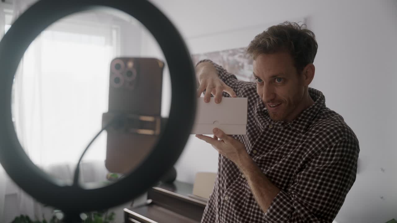 Influencer di social media e creatore di contenuti che recensisce un prodotto o fa un video di unboxing. Parla a uno smartphone che lo riprende con un supporto per ring light in un appartamento o studio casalingo. Entusiasta e felice mentre filma il video