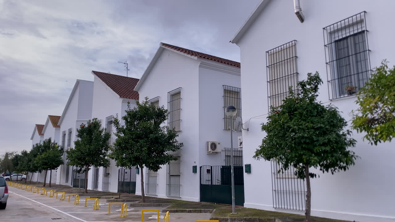 Fila de casas blancas típicas con tejados de tejas bordeando una calle residencial durante un día nublado, con rejas de seguridad y pequeños árboles.