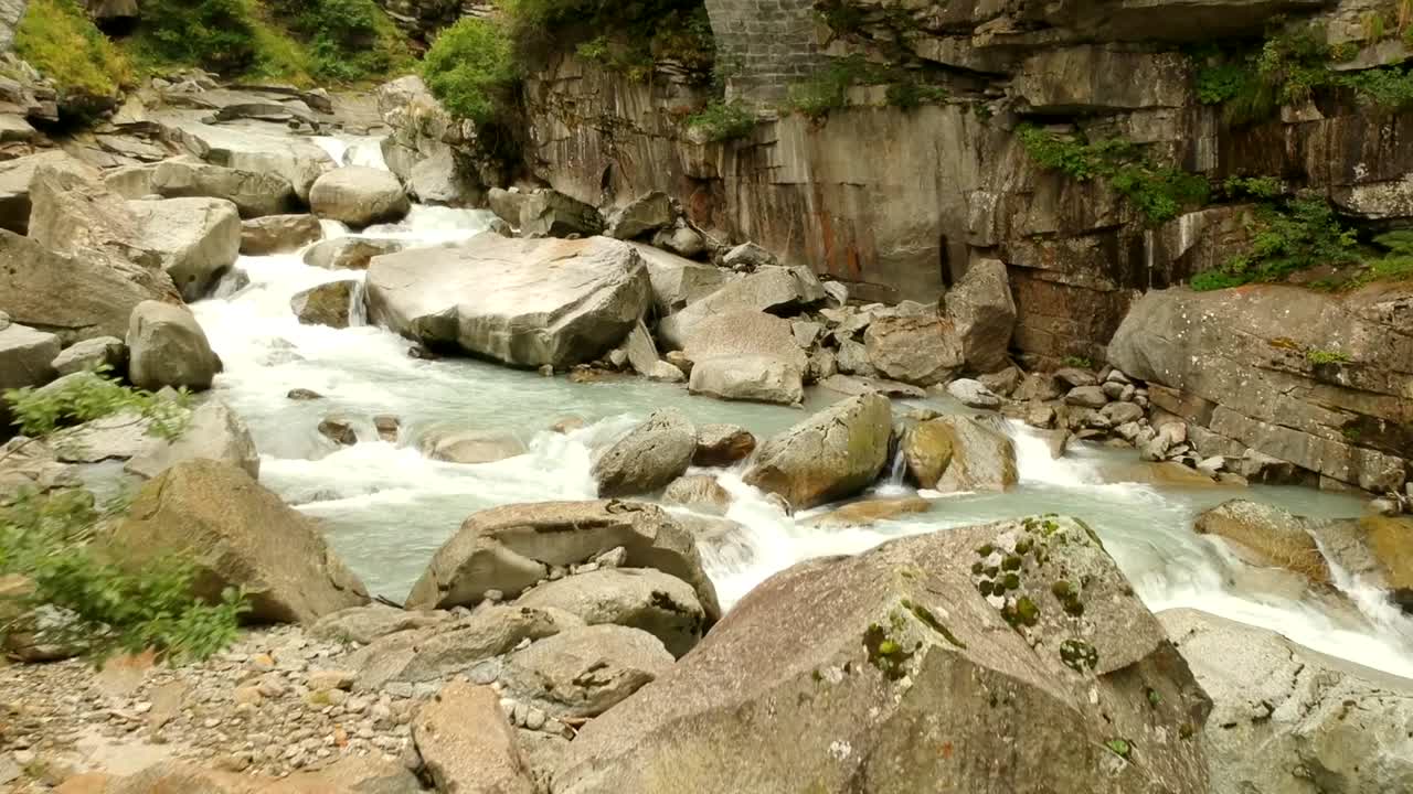 Slow aerial drone shot over a rocky alpine stream with glacial water crashing over lightly coloured, heavily eroded rocks. Approaches a stone bridge at the end. Part two of a series of twelve.
