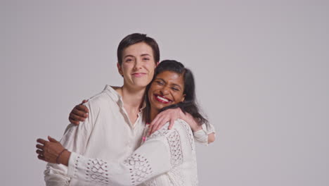 Studio-Portrait-Of-Two-Smiling-Women-Of-Different-Ages-Wearing-Pink-Breast-Cancer-Awareness-Ribbons-Hugging-Against-White-Background