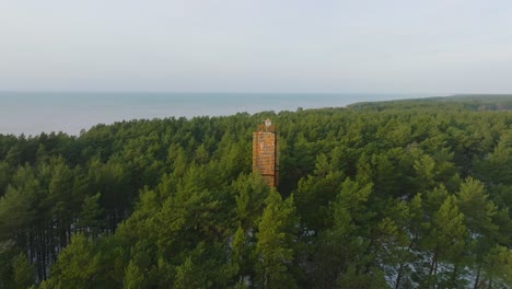 aerial view of bernati lighthouse surrounded by lush green pine tree forest with light snow, nordic woodland, baltic sea coast, sunny winter day, latvia, wide drone shot moving forward, tilt down