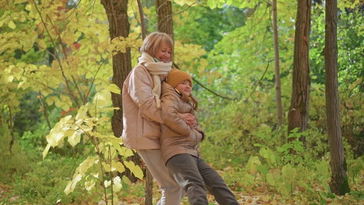 happy mother spinning daughter around in vibrant autumn woodland, both laughing under golden foliage, wearing warm jackets and beanies, surrounded by swirling leaves and dappled sunlight