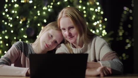 mom and daughter are looking at the laptop screen together. sitting in front of the blurred lights of the christmas tree