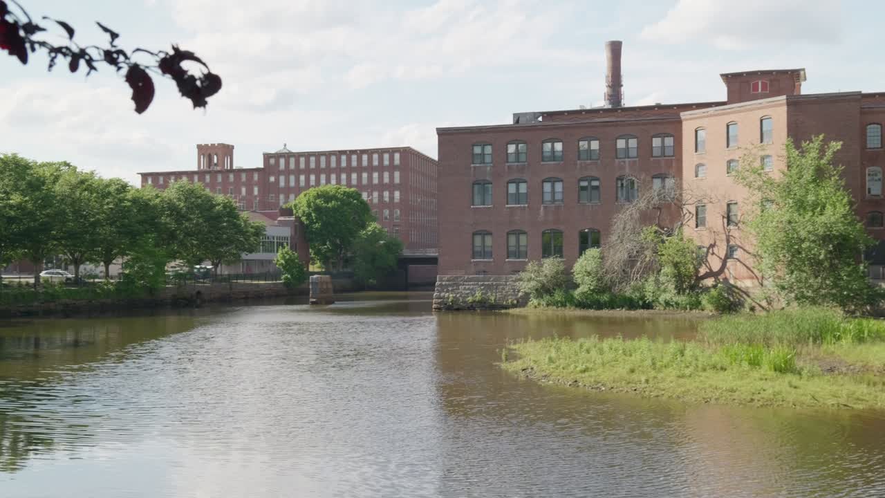 Langsamer Schwenk nach rechts von einem überhängenden Ast, der einen Fluss und ein historisches rotes Mühlengebäude in Dover, NH, USA, enthüllt. Es ist ein heller Sommertag mit blauem Himmel und Wolken