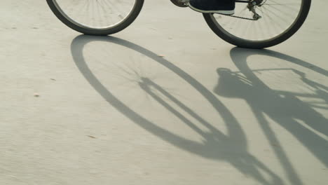 leg view of someone wearing sneakers and black trousers, pedaling a bicycle slowly with a distinct shadow of the bike cast on the sunlit road