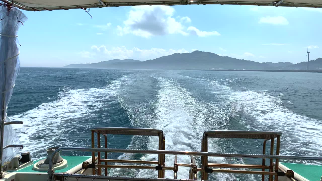 Motor boat at full speed leaving wake of white foam while sailing in the ocean during the day with an impressive mountain in the background seen from the stern of the ship