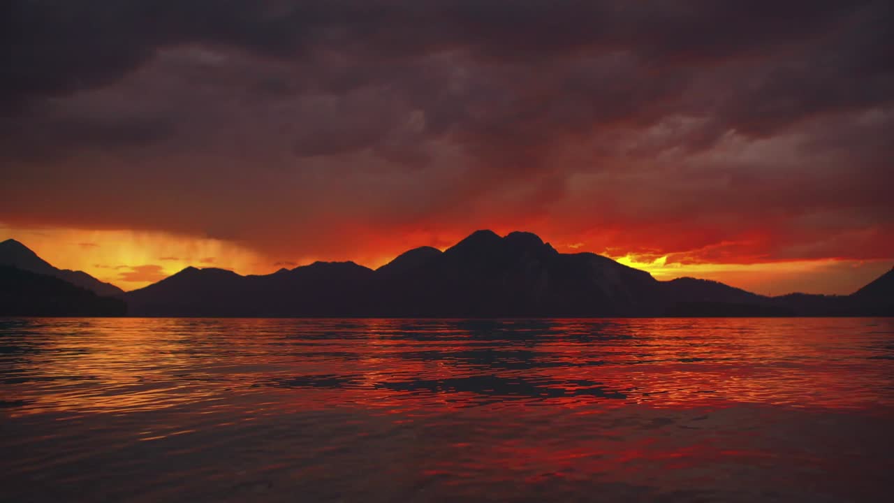 Malerisches Zeitraffer-Cinemagraph / nahtlose Videoschleife eines wunderschönen und romantischen Sonnenuntergangs am natürlichen Bergsee Walchensee in Süddeutschland / Bayern mit sich bewegenden roten Wolken und ruhigen Wellen an einem berühmten Touristen-Urlaubsort. Keine Personen.