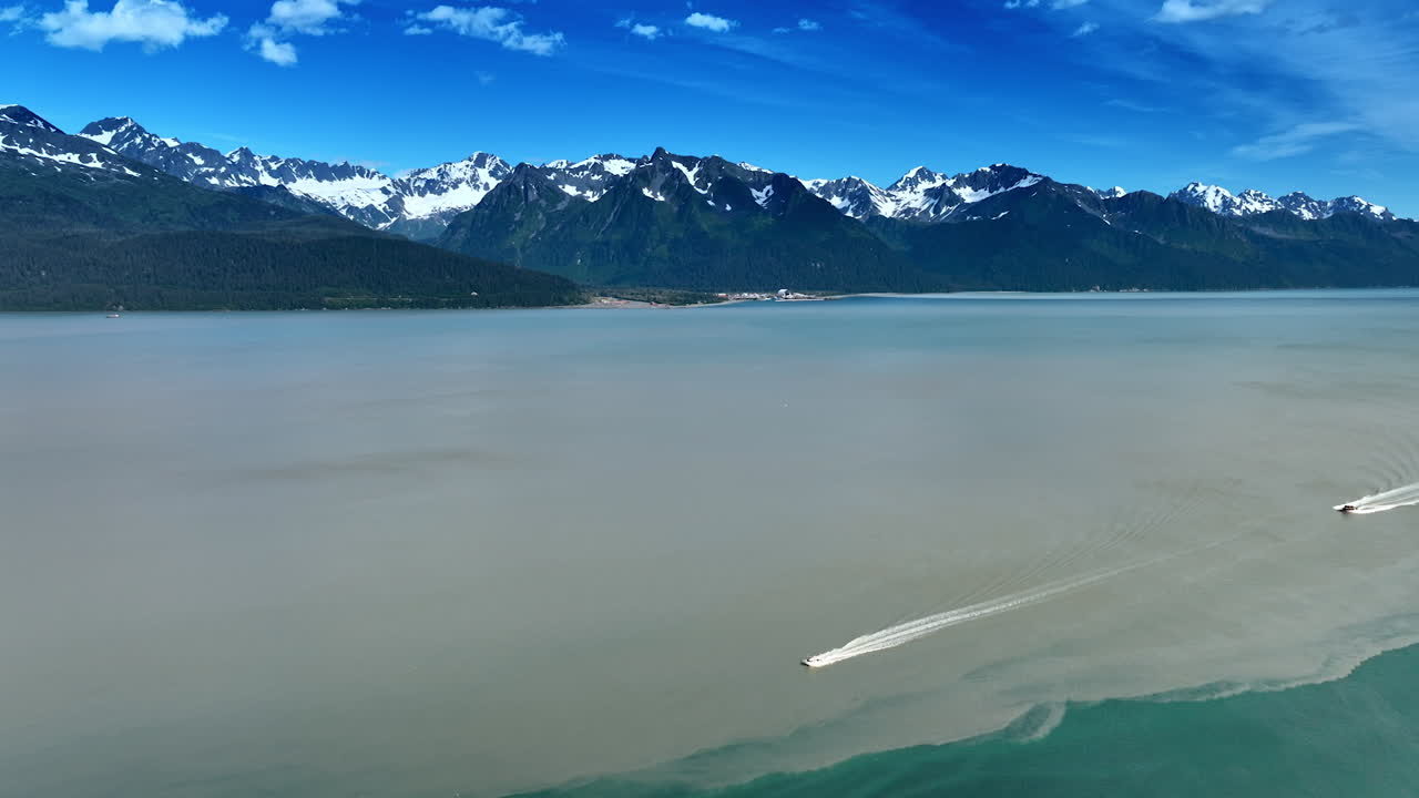 Two speed boats move by the waterscape of a huge lake. Snowcapped mountain range at backdrop. Alaska wilderness. Aerial view