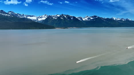 Two speed boats move by the waterscape of a huge lake. Snowcapped mountain range at backdrop. Alaska wilderness. Aerial view