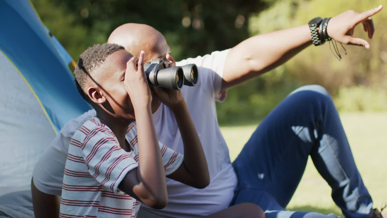 Father guiding son in education as boy reaching for binoculars, teaching birdwatching beside tent