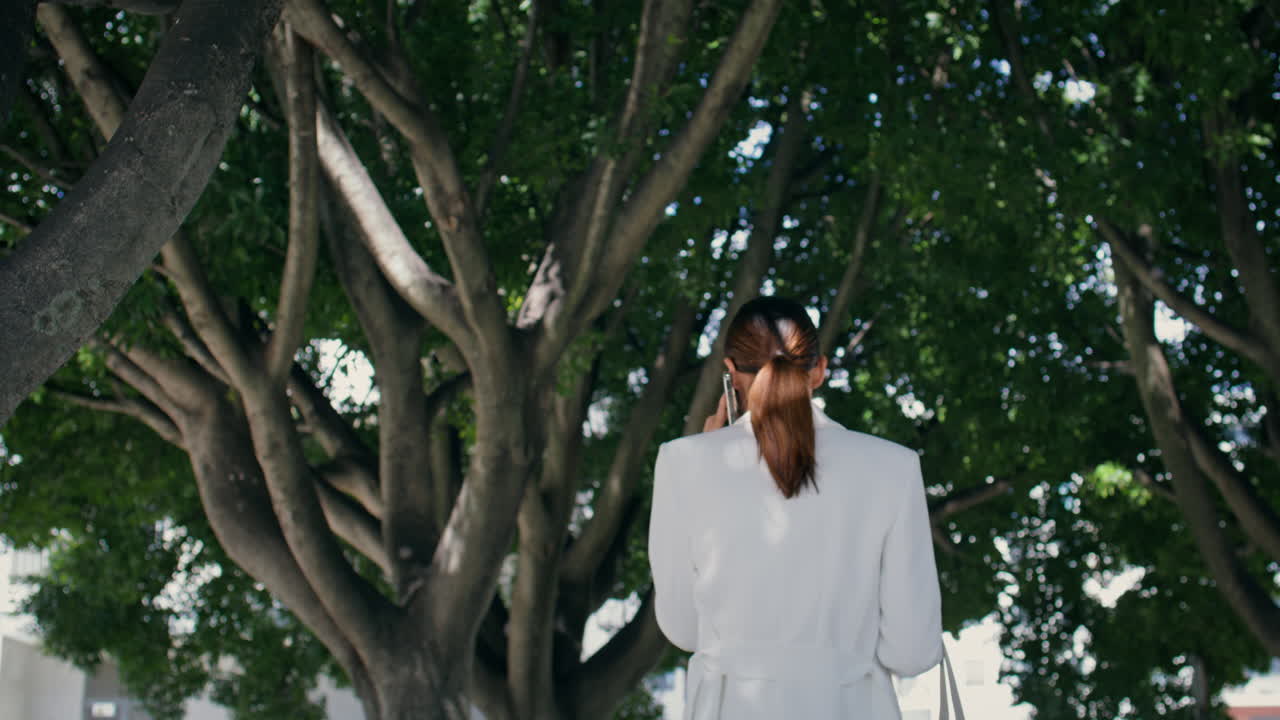 우아한 여자가 휴대전화에 서서 얘기하는 나무. 여자가 전화로 일을 논의하는.