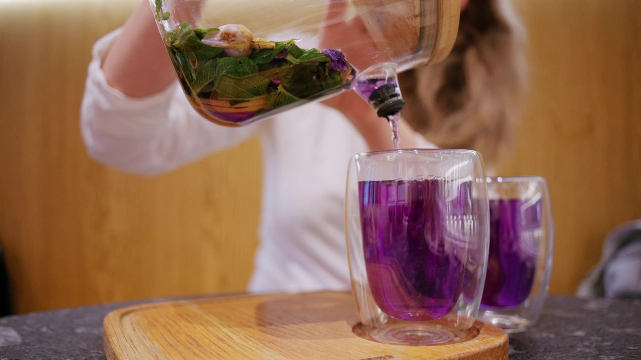 Un'infusione di erbe di un viola vivace, contenente fiori ed erbe fresche, viene versata in una tazza di vetro trasparente.