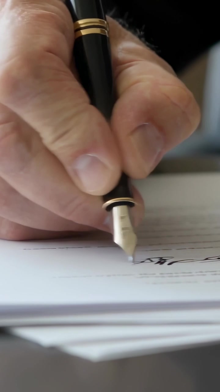 Video verticale: Mano destra che firma sulla linea designata di una scrivania, con penna stilografica e giacca scura.