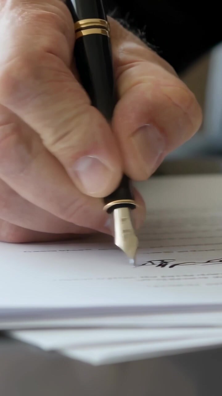 Video verticale: Mano destra che firma sulla linea designata di una scrivania, con penna stilografica e giacca scura.