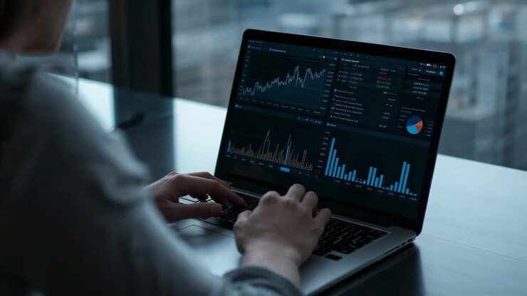 Analytiker i grå skjorte som ser skiftende beregninger, skriver og oppdaterer diagrammer på laptop ved kontorpulten
