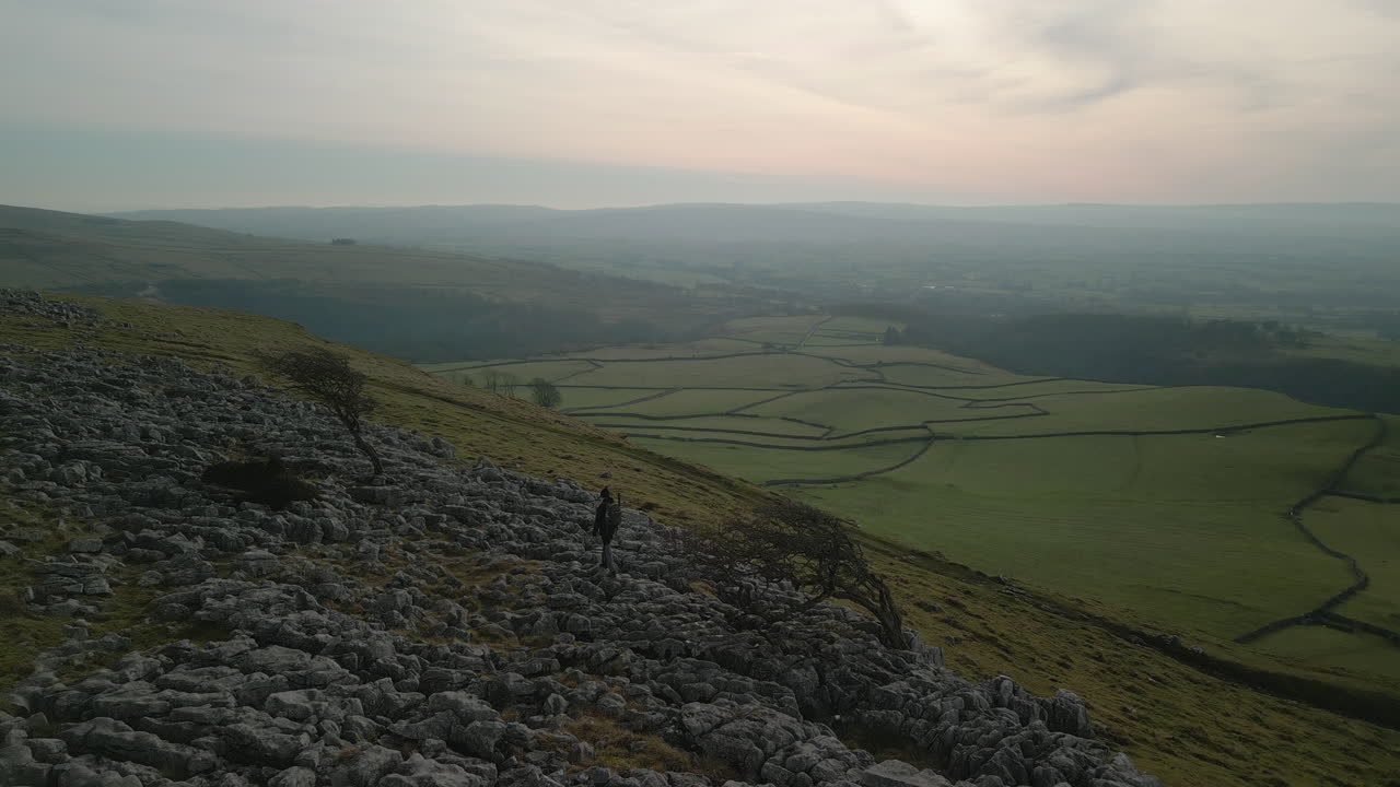 Birleşik Krallık, Yorkshire, Ingleton'daki İngiliz kırsalının yamalı yeşil tarlalarına doğru kayalık bir yamaçtaki yürüyüşçünün üzerinden uçarken.