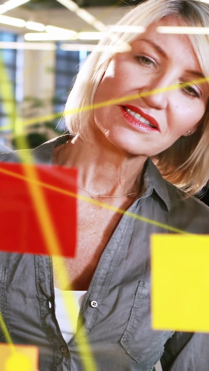 Video verticale: Donna che posiziona una nota rossa, disegna linee gialle su una lavagna di vetro, pianificando la strategia del team.