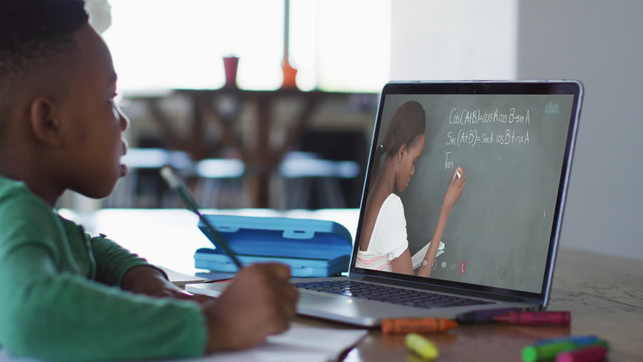 African american boy doing homework while having a video call with female teacher on laptop at home