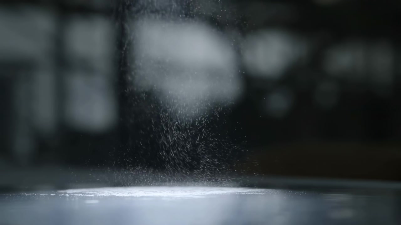 Vista ravvicinata di polvere fine o particelle granulari che scendono in un sottile flusso verticale su una superficie liscia e riflettente, creando una morbida dispersione, un sottile accumulo, un'atmosfera fredda e a bassa luminosità, e
