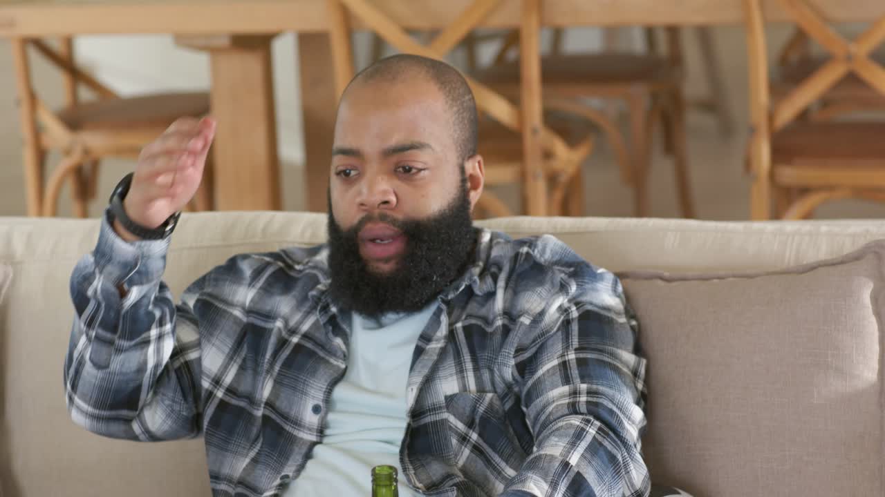 African American man sitting on couch holding beer bottle leaning on soccer ball rubbing forehead