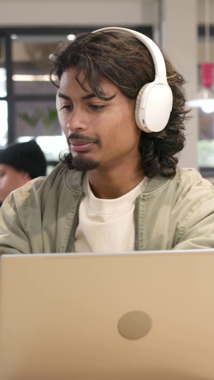 Vertical video: Reaching for headphones, man in green jacket donning them at cafe, for laptop work