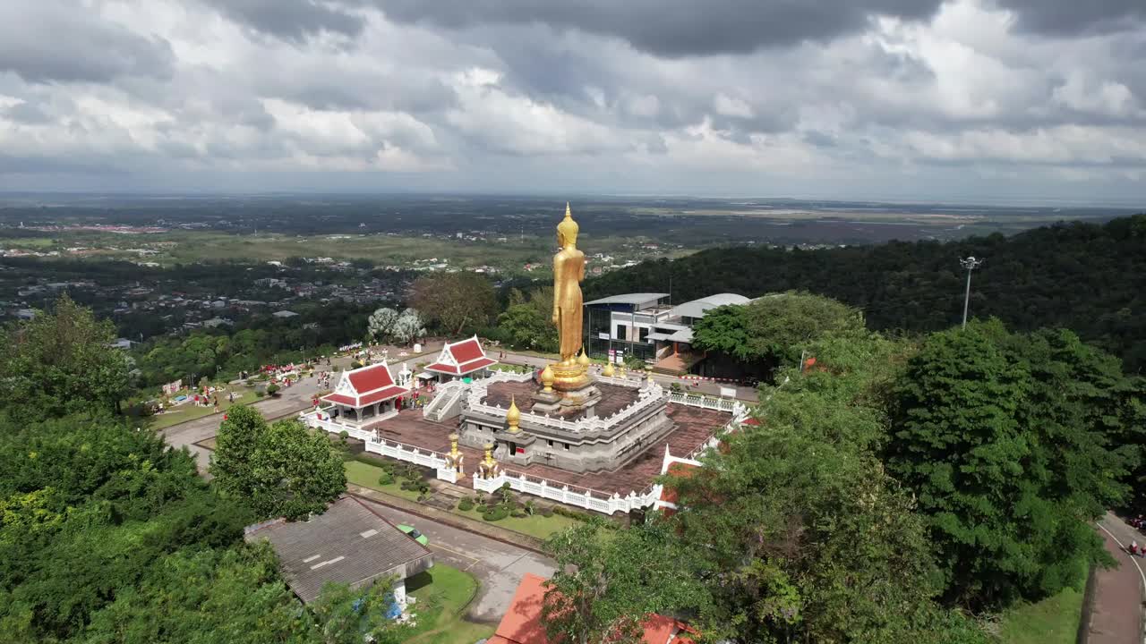 Drone shot of Phetchabun's hilltop temple boasts a towering golden Buddha, red-roofed structures, and lush greenery under dramatic clouds. Captures panoramic views and cultural majesty