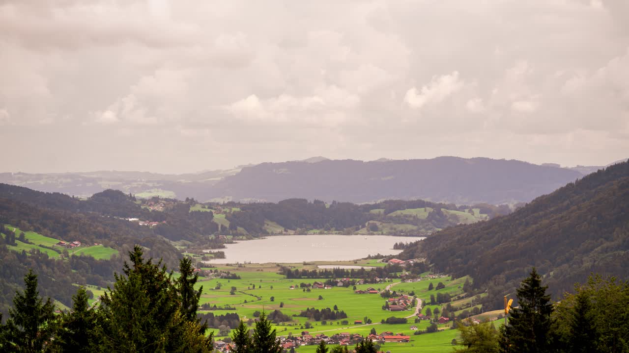 This timelapse video captures the tranquil Alpsee nestled in the hills of Allgäu, Bavaria, Germany. Sailboats drift across the water, surrounded by rolling pastures and forests beneath a cloudy sky.