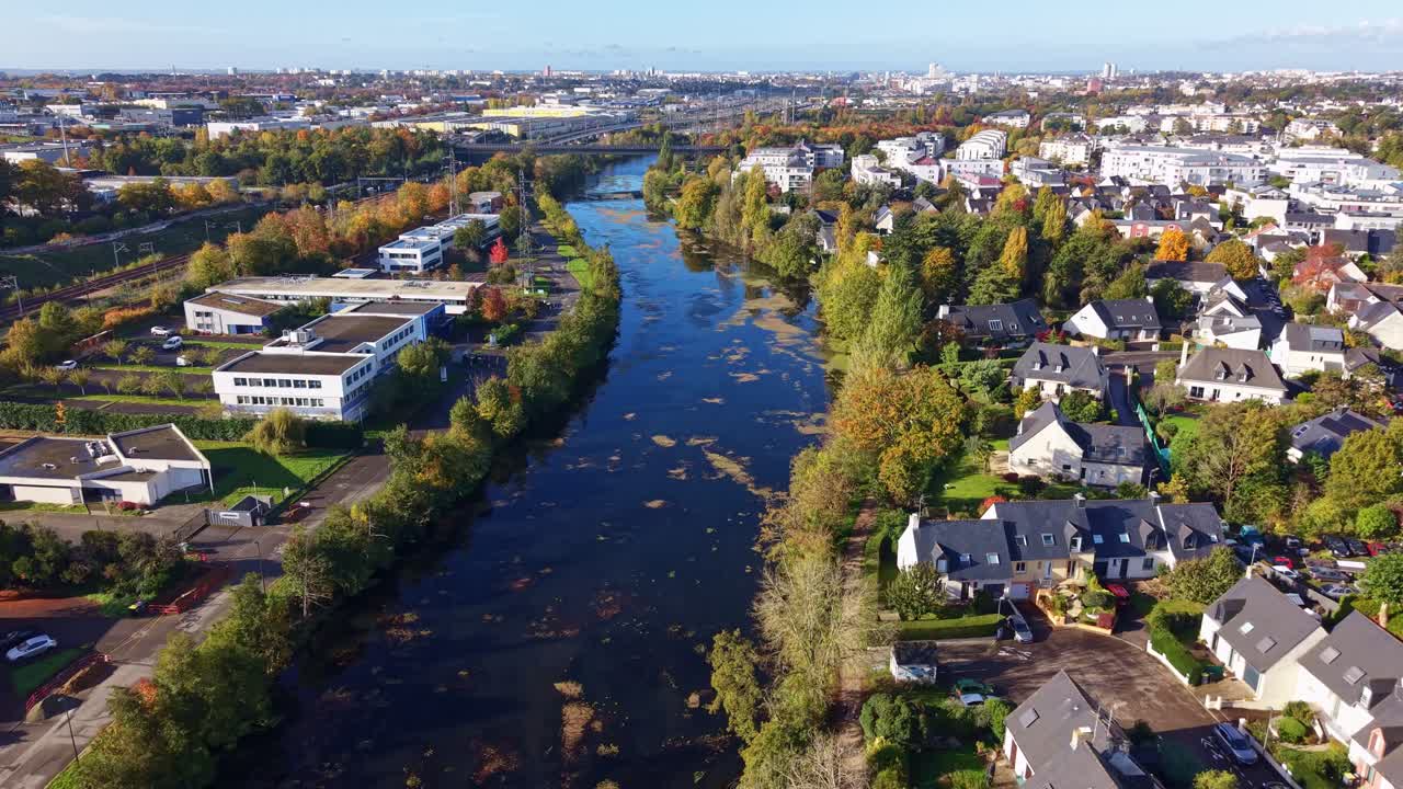 La Vilaine river, framed by suburban homes and autumn trees, flows through Cesson-Sévigné in Brittany, seen by drone pulling out, with Rennes skyline in the distance,