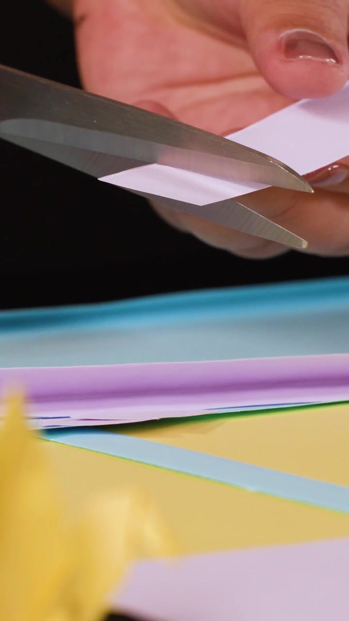A person uses metal scissors to cut small purple paper strips over a colorful surface