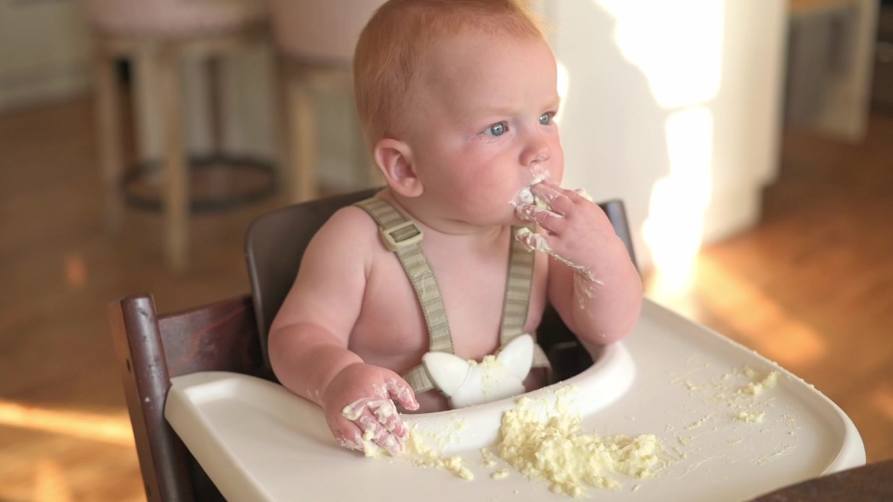 Baby with light red hair sits in high chair, engaging with soft food using hands, creating a playful mess on the tray and floor, surrounded by a warm, inviting kitchen atmosphere