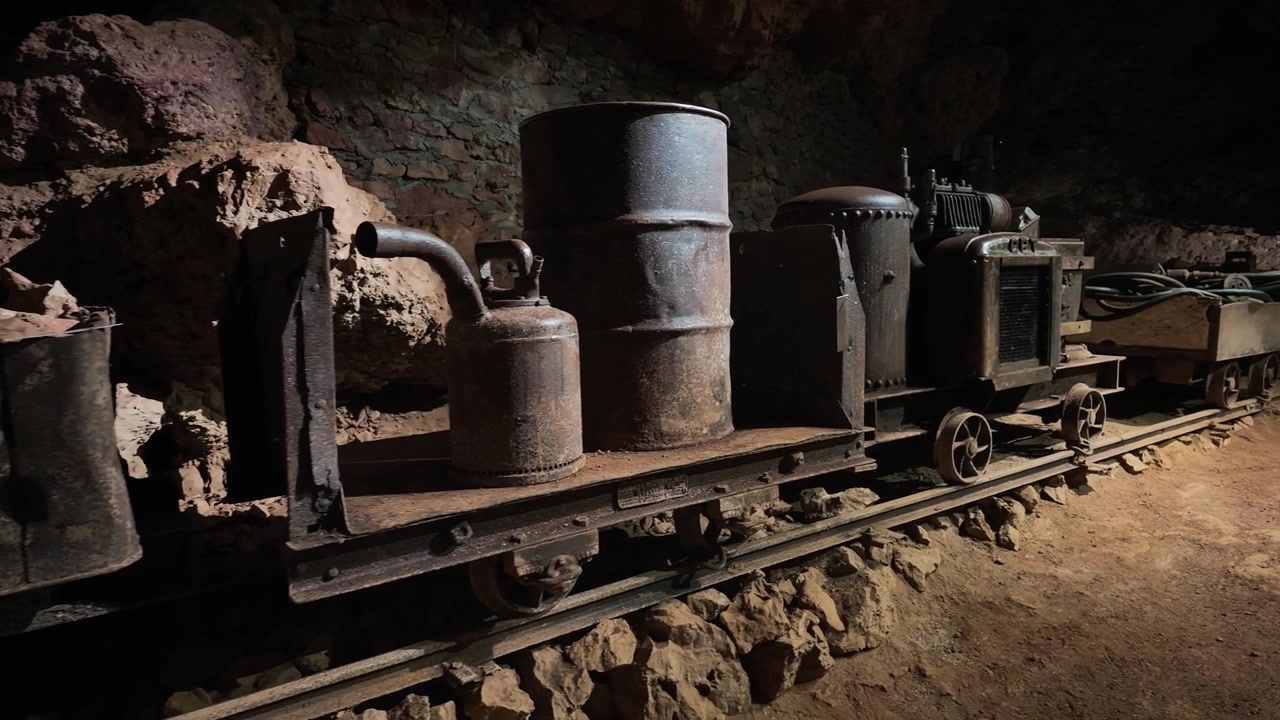 Old mine cart loaded with a rusted barrel and metal equipment on narrow tracks inside a dim underground tunnel.