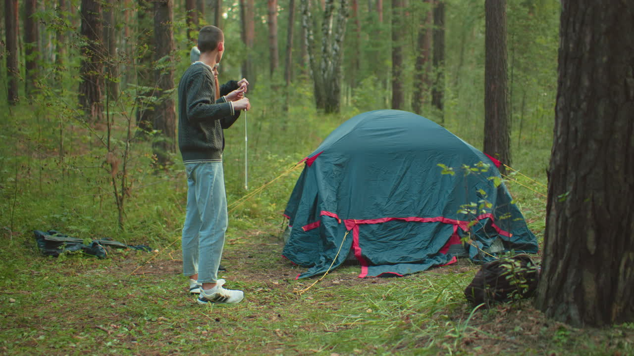Couple working together to fix flexible tent pole beside pitched tent in forest setting, surrounded by trees and grass, wearing casual outfits and engaging in collaborative outdoor camping activity