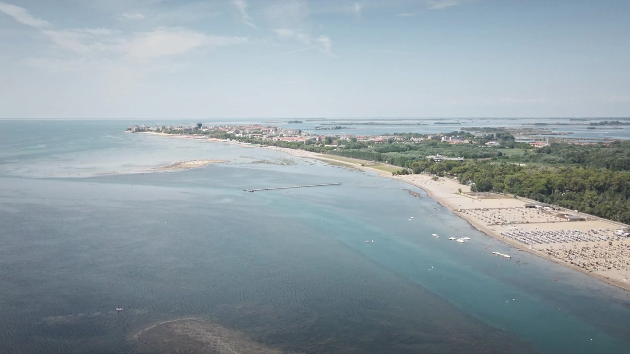 Drone 4K UHD vola fluidamente in avanti sopra le acque basse e calme della laguna di Grado, catturando la vasta costa, spiagge sabbiose punteggiate di ombrelloni e lussureggianti pinete verdi.