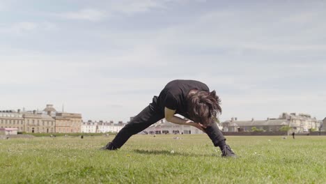 Young-man-in-black-stands-on-grass-near-buildings-outside-and-stretches