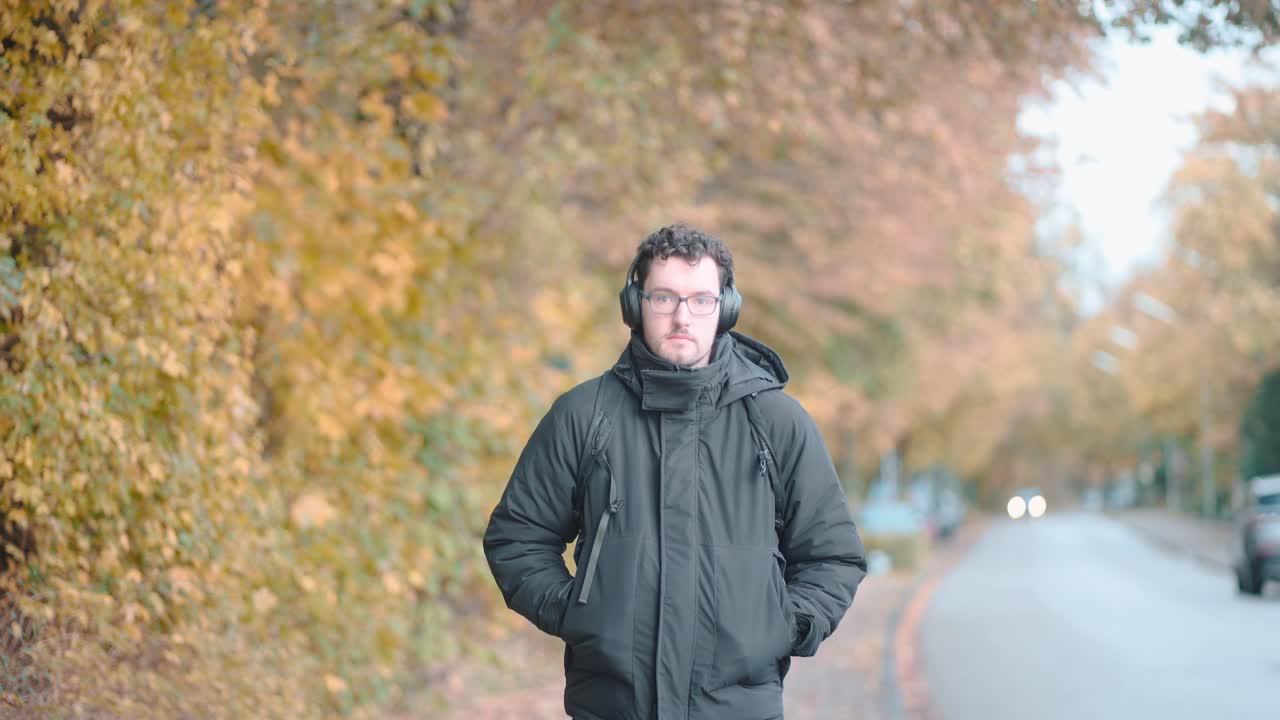 수염과 안경을 쓴 세련된 유럽인 남자가  ⁇ 니히의 가을 교외를 여유롭게 산책하고, 헤드폰에서 멜로디에 깊이 몰입하고, 계절의 평온을 흡수합니다.