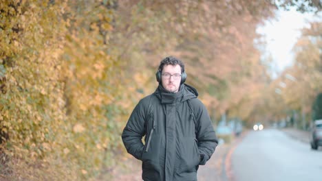 Stylish-European-man-with-a-beard-and-glasses-leisurely-strolls-the-autumnal-suburbs-of-Munich,-deeply-immersed-in-melodies-from-his-headphones,-absorbing-the-seasonal-tranquility