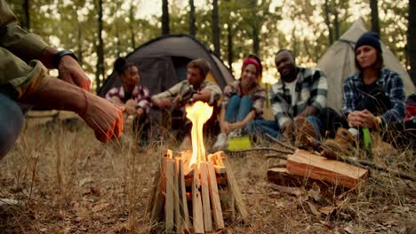 a small fire burns during a halt near a group of people on a hike in special hiking clothes against the backdrop of tents in a green autumn forest with withered grass. a man sits near a fire and warms his hands during a stop on a hike