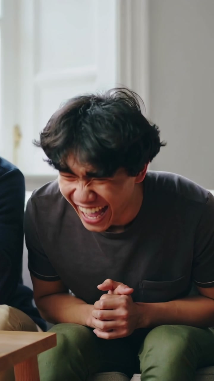 Vertical video: Smiling Asian male bursting out laughing during chat on couch, wearing gray tee