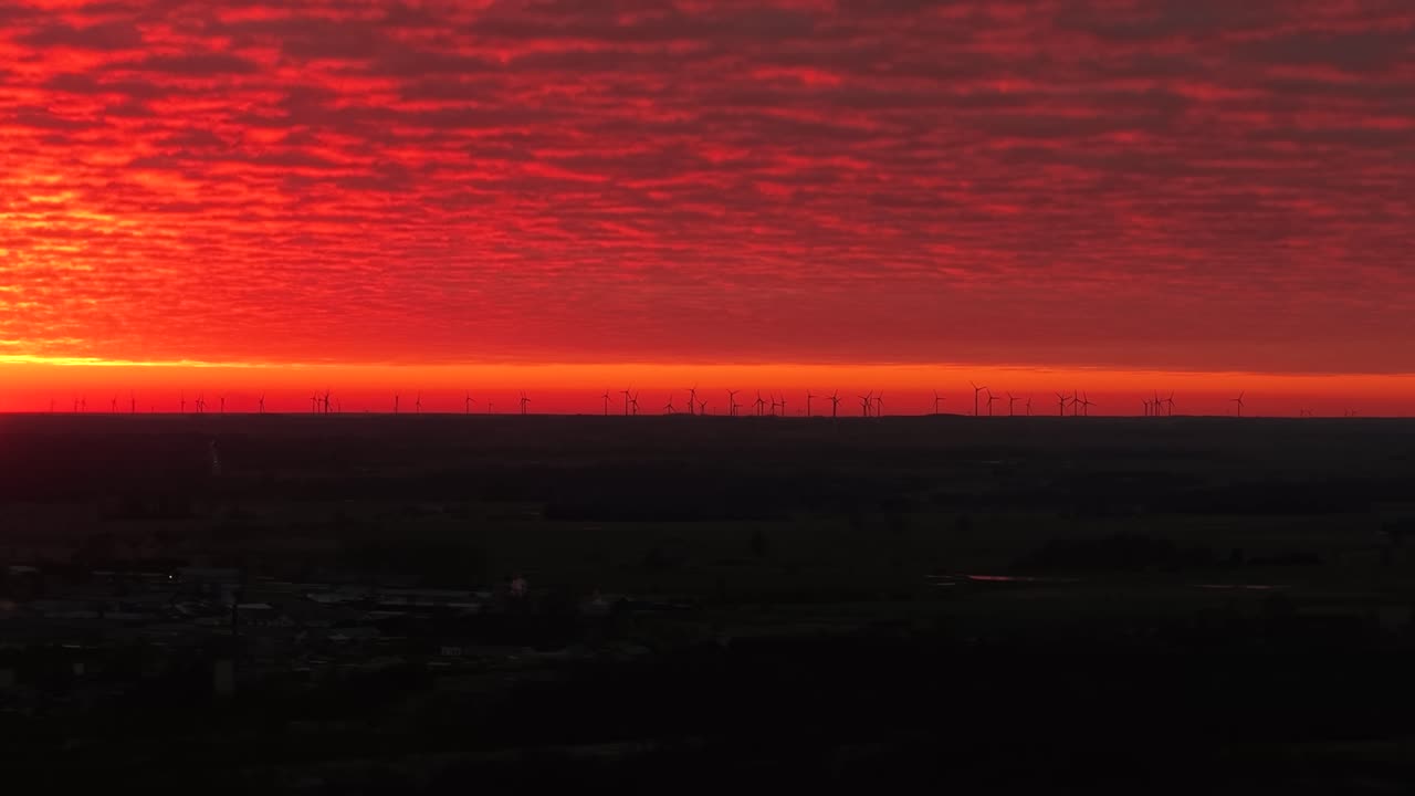Drohnenvideo, das einen markanten Sonnenuntergang über einem Windpark einfängt. Der leuchtend rote Himmel kontrastiert mit den silhouettierten Turbinen und schafft so eine atemberaubende, ruhige Landschaft. Perfekt für Energiethemen.
