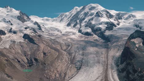 Tilt up shot showing the Gorner Glacier surrounded by snow-covered peaks in the Alps. The movement reveals the immense ice flow, rock formations, and alpine environment beneath a clear blue sky