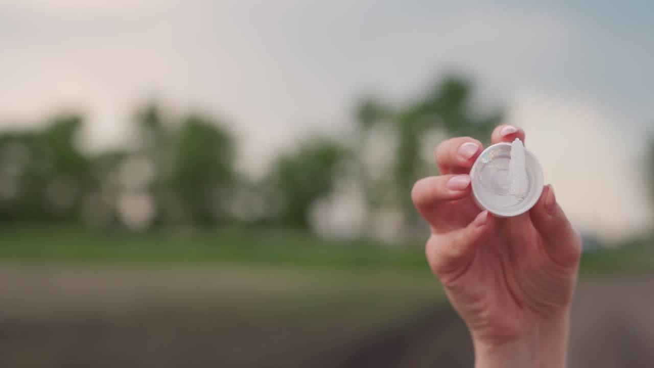 close up of woman hand holding bubble wand container filled with soap solution shimmering in sunlight against blurred farmland and dirt road background joyful bubble