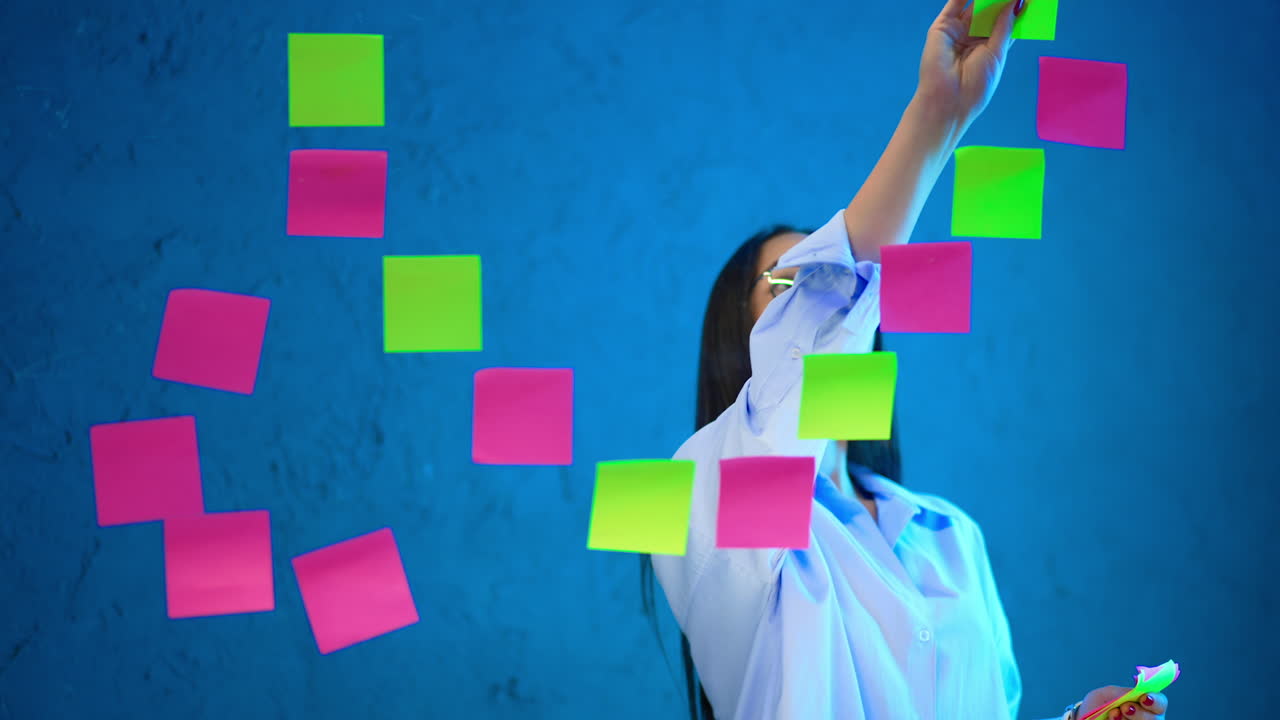Long-haired Caucasian lady wearing glasses and shirt puts stickers on the glass board. Sorting out plans and ideas. Brainstorming concept.