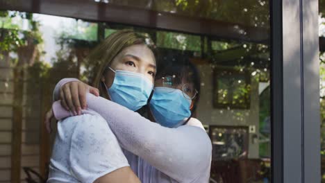 madre y hija asiáticas mirando a través de la ventana con máscaras faciales abrazando