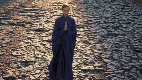 young monk wearing a kasaya, praying barefoot on an ancient stone road at dawn. hands joined in prayer, embodying tranquility and devotion in the soft light of sunrise