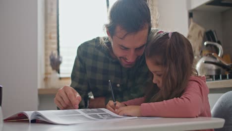 Happy-father,-brunette-man-in-a-checkered-green-shirt-teaches-his-little-brunette-daughter-to-write-while-sitting-at-the-kitchen-table-in-the-kitchen-during-the-day-in-a-modern-apartment