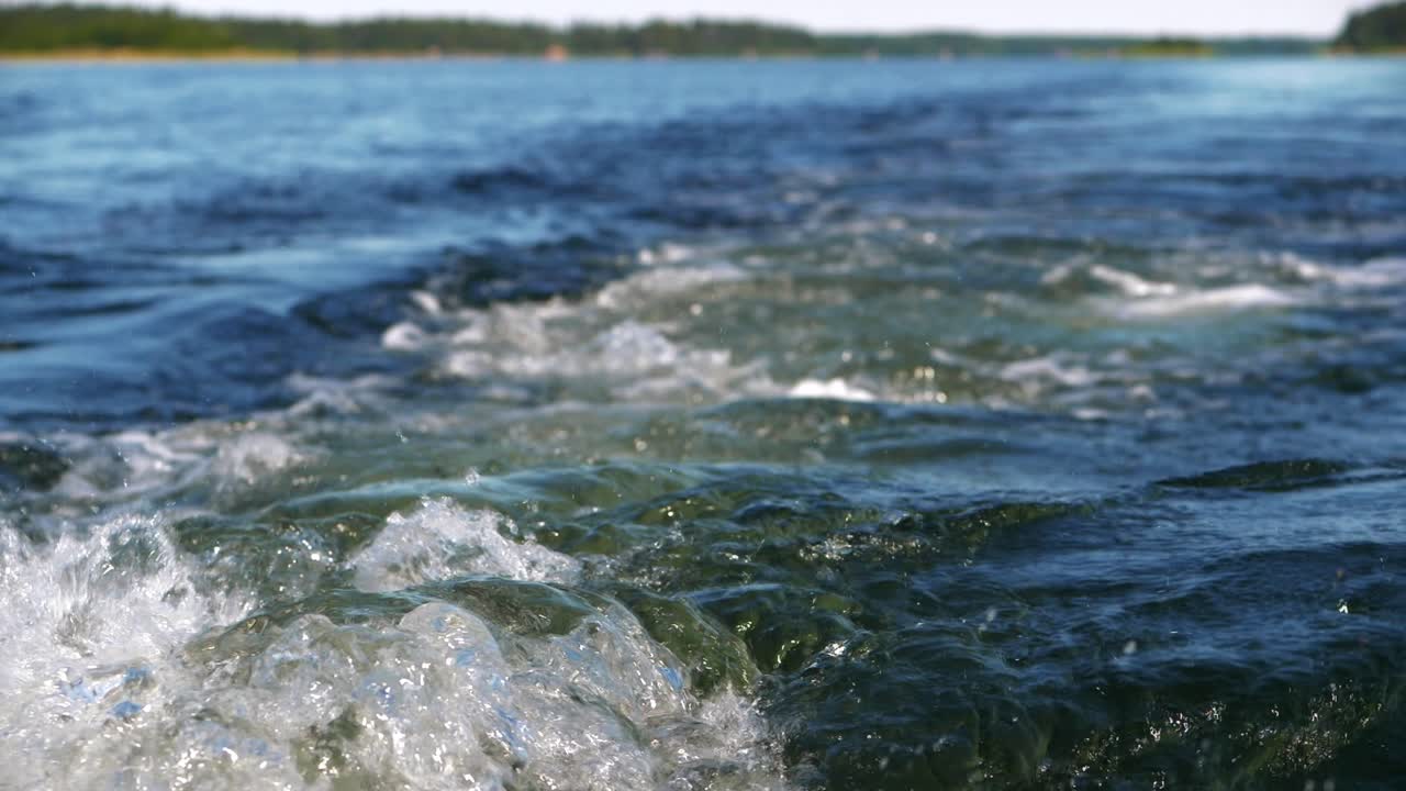 Bådmotorens skumspor i vandet i slowmotion med kysten i baggrunden.