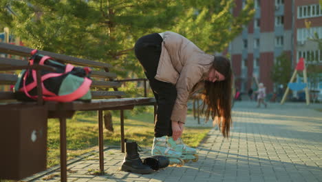 a young woman in a peach jacket and black trousers is bending over to fasten her rollerblades, preparing for skating. a bag is on the bench beside her. children playing in the background