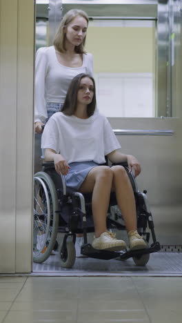 blonde woman pushes sister with disability in wheelchair after door of modern elevator opens. ladies go to therapist appointment in hospital