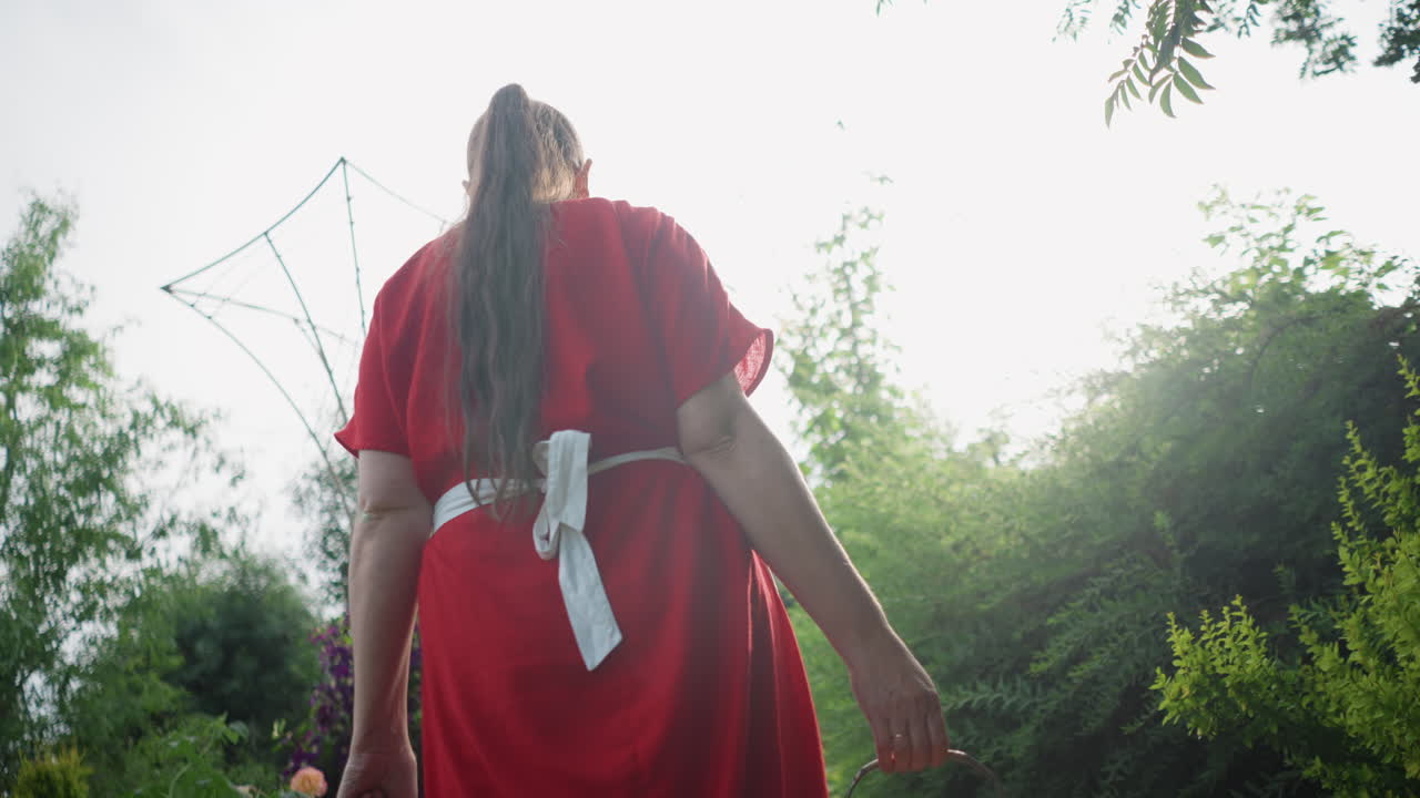 Mulher branca com vestido vermelho carregando uma cesta trançada ao longo de um caminho de jardim ensolarado, a vista em ângulo baixo enfatiza a saia esvoaçante e a trança, avental branco, com uma atmosfera de oferta ritualística, folhagem vibrante e contraluz brilhante.